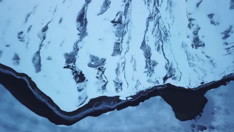 Scenic-Drone-view-Frozen-mountain-and-river-flowing-alongside-iced-lake-in-Switzerland