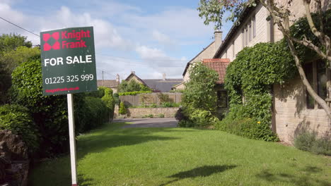 Static-Shot-of-British-‘For-Sale’-Real-Estate-Sign-on-Sunny-Summer’s-Day-with-Rural-Village-Home-in-Background-in-Slow-Motion