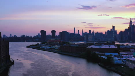 aerial-drone-footage-pull-away-over-purple-water-from-the-refection-of-the-purple-sky-sunset,-as-camera-pulls-straight-back-from-New-York-City-skyline-view