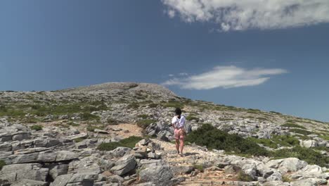 Girl-in-summer-hiking-outfit-walking-up-mountain-in-arrid,-mediterranean-landscape