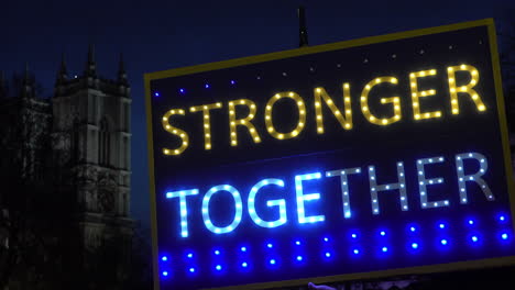 A-passenger-plane-passes-behind-an-illuminated-placard-that-says-“Stronger-Together”-and-Westminster-Abbey-is-seen-in-the-background-at-night