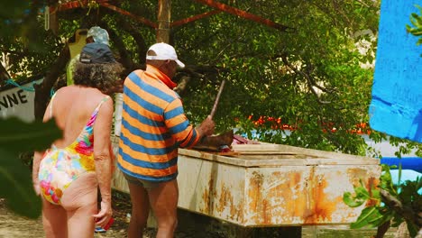 Local-Fisherman-Cutting-The-Fresh-Caught-Swordfish-Using-A-Big-Knife-While-The-Female-Tourist-Is-Watching-On-The-Back-In-Curacao