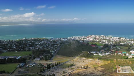 Panning-shot-of-coastal-town-next-to-bay,-aerial-view-from-mountain,-Hermanus,-South-Africa