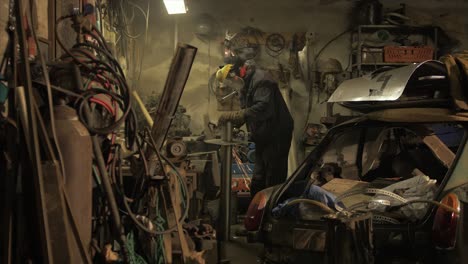 Man-angel-grinds-metal-plate-on-steel-piping-in-messy-workshop