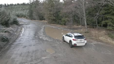 A-lone-white-Subaru-Crosstrek-dives-into-a-deep-mud-puddle,-slow-motion-aerial,-illustrative-editorial