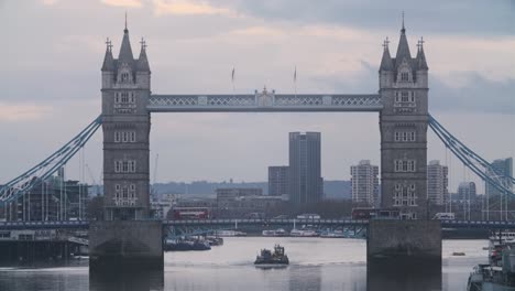 Traffic-London-bus-passing-over-boat-under-Tower-Bridge-London-on-a-cloudy-evening-telephoto