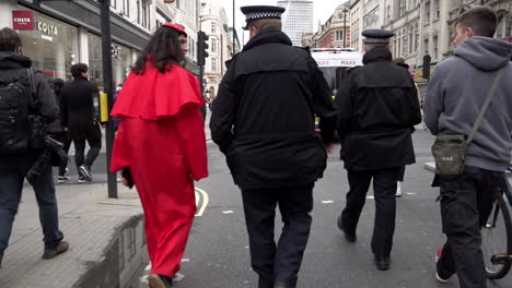 Slow-motion-of-a-man-dressed-in-a-red-Catholic-Cardinal-smock-walking-along-side-a-police-officer-on-a-Coronavirus-and-QAnon-conspiracy-protest