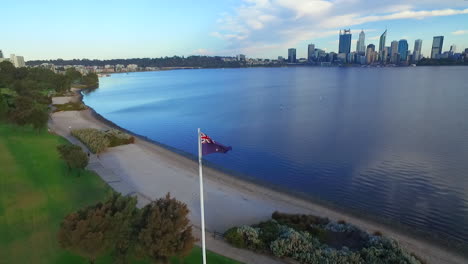 Aerial-tilt-down-flyover-of-Australian-flag-pole-in-park,-city-and-river-in-background