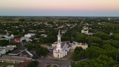 Dolly-out-of-neo-gothic-church-surrounded-by-trees-in-idyllic-Santa-Elisa-countryside-town-at-sunset-with-farms-in-background,-Entre-Rios,-Argentina