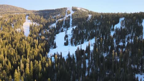 Drone-birdseye-wideangle-footage-over-ski-resort-on-a-bright-cloudless-day-in-Lake-Tahoe,-USA,-showing-skiers-down-a-slope,-chairlifts-moving-up-the-mountain-and-the-surrounding-scenic-alpine-forest