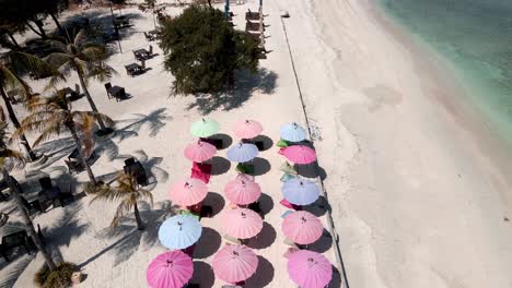 Aerial-fly-over-multicolored-umbrellas-on-golden-beach-beside-tropical-ocean-shore-on-Gili-Trawangan