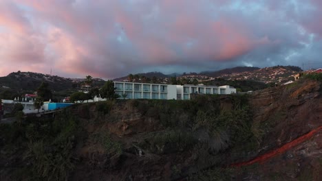 Drone-shot-moving-upwards-and-tilting-down-on-the-luxury-hotel-on-the-cliff-of-Ponta-do-Sol-in-Madeira-during-sunset-with-dramatic-storm-clouds-in-the-background