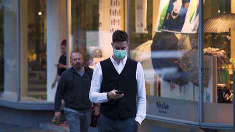 An-elegant-young-guy-wearing-a-green-protective-Covid-19-facemask,-a-black-waistcoat,-white-shirt-and-jeans-crossing-the-street-and-calling,-heading-towards-the-camera,-a-shop-in-the-background,-4k