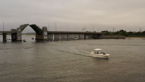 A-low-angle-aerial-view-of-a-small-boat-sailing-on-Baldwin-Bay,-past-a-draw-bridge-opening-on-a-cloudy-day