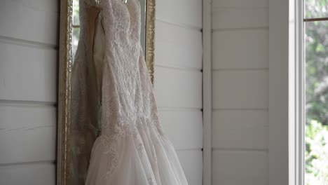 View-from-the-side-of-a-wedding-gown-hanging-in-a-well-lit-clean-white-room-in-front-of-a-mirror-with-a-beautiful-golden-frame