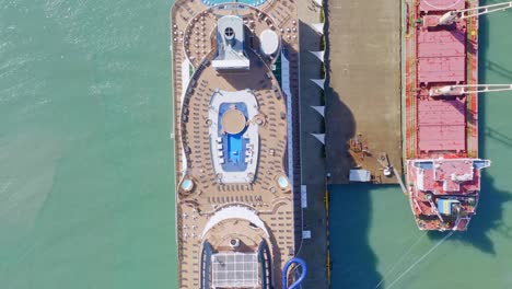 Aerial-top-down-shot-of-luxury-cruise-ship-with-private-swimming-pool-and-chute-beside-docking-industrial-ship