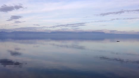 Isolated-fisherman-boat-in-silent-waters-of-lake-reflecting-the-cloudy-sky-and-small-island-in-middle