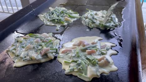 Cooking-Korean-seafood-scallion-pancakes,-shaping-wet-ingredients-into-flat-symmetrical-circles-over-a-super-hot-flat-iron-grill