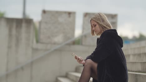 Attractive-young-blond-girl-sitting-on-stairs-outdoors-during-windy-day-and-watching-smartphone