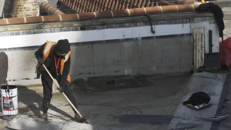 Männlicher-Arbeiter,-Der-Bitumenmastix-Auf-Die-Dachwohnung-Eines-Gebäudes-In-London-Aufträgt