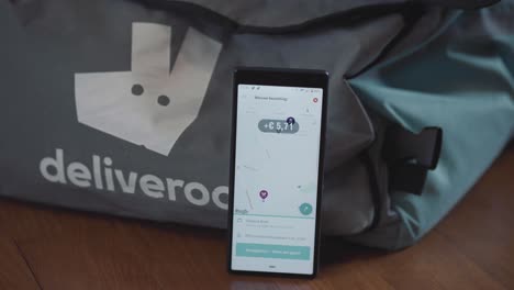 Deliveroo-Order-Coming-In-For-Rider-Using-Map-On-Smartphone-Leaning-On-Delivery-Bag-On-The-Table