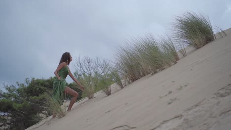 Dark-haired-women-in-a-smaragd-green-calssy-elegant-dress-walkup-upwards-a-sand-dune-hill-with-a-cloudy-grey-blueish-sky---dramatic-cinematic-slowmotion