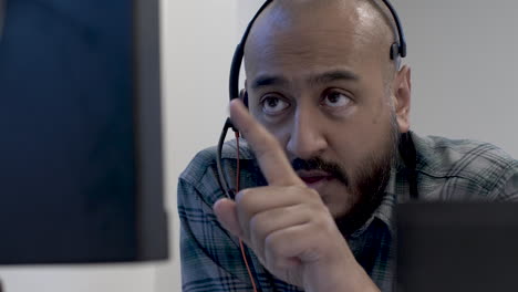 Bald-UK-Asian-Male-Talking-To-Colleague-Wearing-Headset-Microphone-Viewed-Behind-Monitor