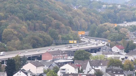 Wide-shot-of-traffic-on-german-autobahn-highway-with-many-cars-and-trucks-beside-green-mountain-and-housing-area