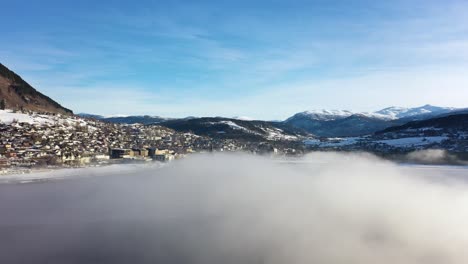 Voss-morning-aerial-flying-through-frost-haze-above-Vangsvatnet-lake-while-looking-at-town-of-Voss-with-mountains-and-blue-sky-background