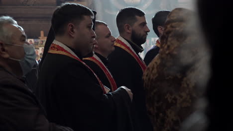 Four-priests-stand-in-line-in-front-of-light-pouring-through-a-church-window-at-the-funeral-of-a-Ukraine-Soldier-during-the-Russian-invasion-of-the-country