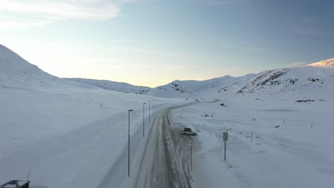 Truck-from-TSL-transport-crossing-Hemsedal-mountain-on-snow-covered-road-at-sunset-hours---Aerial-flying-over-road-with-truck-overtaking-drone-and-revealing-itself-in-middle-of-clip