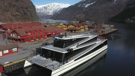 Fully-electric-passenger-catamaran-Future-of-the-fjords-alongside-in-Flam-Norway---Aerial-slowly-approaching-and-flying-over-vessel-with-Flam-village-and-mountain-background