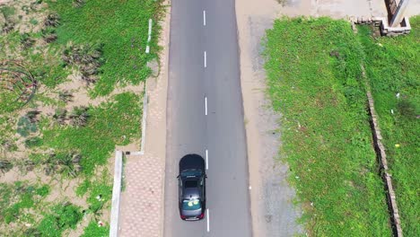 aerial-footage-following-a-luxury-dark-car-with-a-white-car-trailing-it-behind-on-an-empty-concrete-road,-greenery-and-sand-on-both-sides,-road-trip,-outdoors,-sunroof,-coconut-trees,-building,-bright