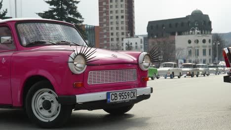 Convoy-of-Eastern-European-Trabant-retro-classic-cars-drive-through-city-streets,-Colourful-pink-car