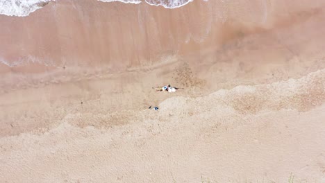 zooming-out-shot-of-a-heterosexual-couple-lying-side-by-side-on-an-empty-sandy-beach-with-gentle-white-foam-waves-lazily-lap-up-the-shore,-greenery-visible,-bright-outdoor-day,-romantic,-intimate-view