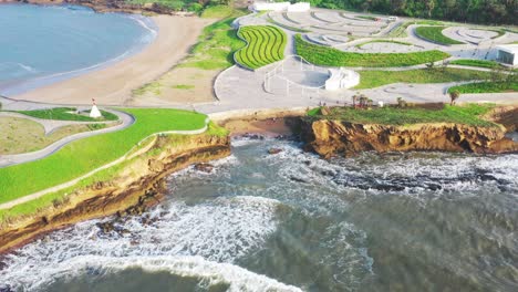 shot-slowly-zooming-out-from-an-expansive-structure-right-by-the-sea,-gentle-foamy-white-waves,-modern-architecture,-bright-green-patches,-bright-sunny-day,-cliffside-visible,-natural-colorful-scenery