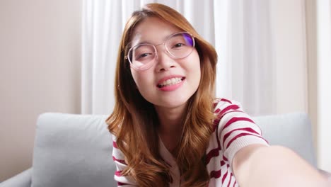 Porträt-Einer-Selfie-Asiatin-Mit-Brille,-Die-Bei-Einem-Online-Videoanruf-Lächelt-Und-Positive-Emotionen-Hervorruft,-Während-Sie-Auf-Dem-Sofa-Im-Wohnzimmer-Sitzt
