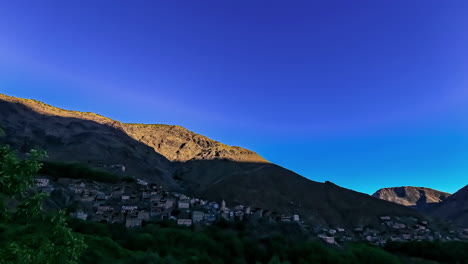 Tiro-De-Lapso-De-Tiempo-De-Hermosas-Colinas-De-Iluminación-Del-Amanecer-Y-Pequeño-Pueblo-En-Marruecos,-Montañas-Del-Atlas---Hermoso-Cielo-Azul-En-El-Fondo