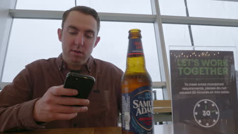 Ein-Kaukasischer-Mann-überprüft-Sein-Mobiltelefon,-Während-Er-In-Einer-Bar-Am-Internationalen-Flughafen-Von-Seattle-Tacoma-Bier-Trinkt