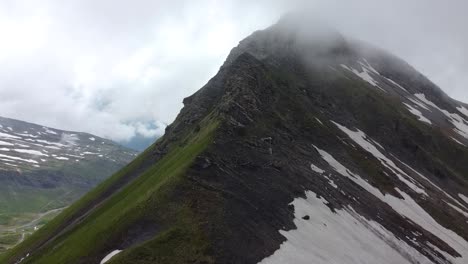 Drone-shot-of-a-Dark-Mountain,-partially-covered-in-snow-and-in-the-Clouds