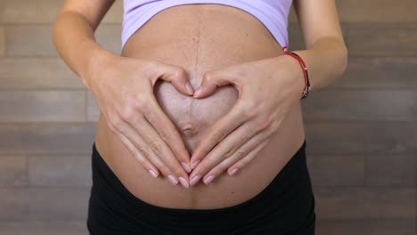 Pregnant-woman-forms-a-heart-with-hands-on-big-belly-button,-close-up