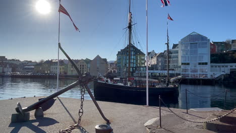 Old-wooden-boat-motoring-out-of-the-Ålesund-Harbor,-past-anchor-and-Molja-LIghthouse,-Thon-Hotel-and-Academy,-bright-sun,-panning-right-to-left