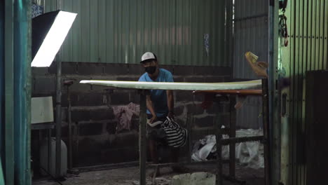 Two-Asian-men-working-on-homemade-DIY-surfboard,-pouring-and-spreading-paint