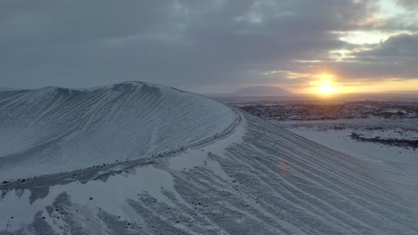 Aerial-backwards-shot-of-snow-covered-Hverfjall-Volcano-Crater-during-sunset-at-horizon---Northern-Iceland,Europe