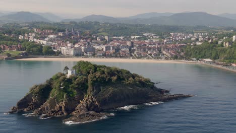Lonely-house-in-small-rocky-island-with-San-Sebastian-city-in-background,-cinematic-aerial-view