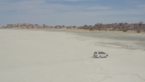 Traveling-SUV-Car-On-Arid-Desert-At-Kubu-Island,-Botswana-With-Baobabs-And-Salt-Pan-In-Background