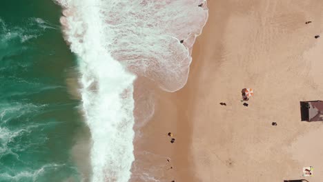 Static-aerial-drone-showing-endless-wild-sea-waves-breaking-on-the-sandy-coast-line-while-people-hang-out-on-the-beach