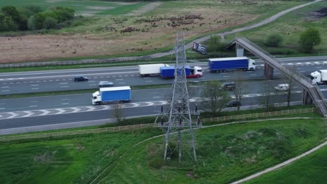 Vehicles-on-M62-motorway-passing-pylon-tower-on-countryside-farmland-fields-aerial-view-high-right-orbit