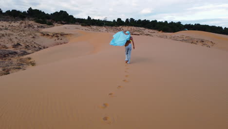 Aerial,-model-with-blue-hijab-walk-along-desert-sand,-waving-silk-fabric-in-wind