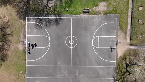 An-aerial-view-over-four-people-playing-basketball-on-a-court-surrounded-by-dry-trees-on-a-sunny-day